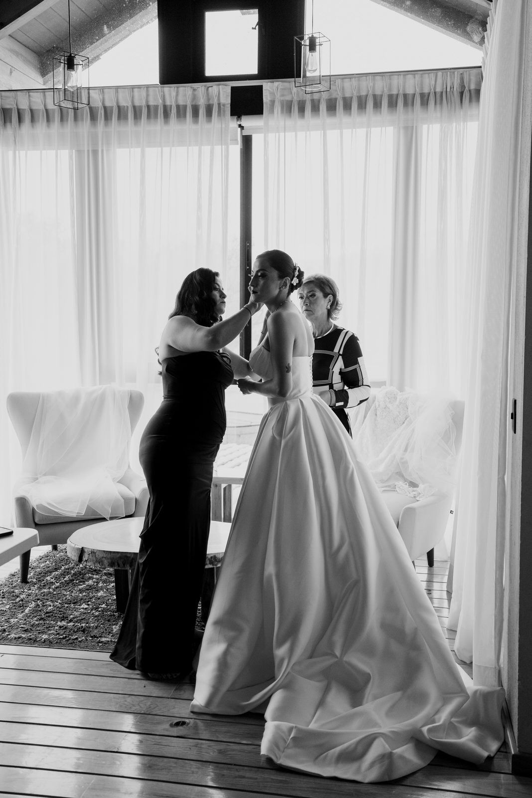A black and white wedding photo showing final dress preparations in a bright room.