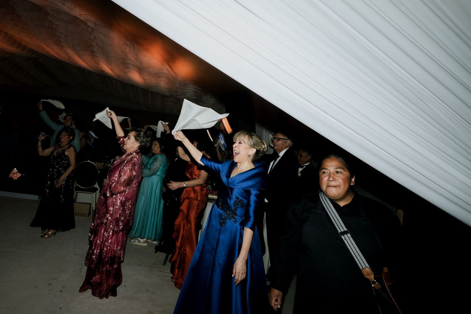 Wedding guests lifting a large white cloth during a festive reception celebration indoors.