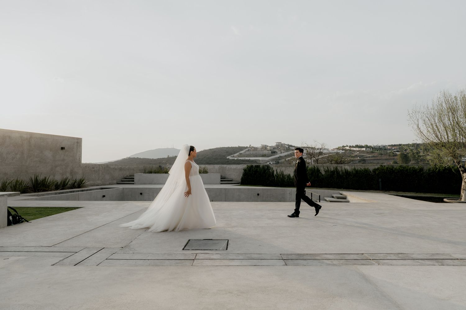 Bride in flowing white gown and groom in black suit walk across modern concrete terrace with scenic hills backdrop.
