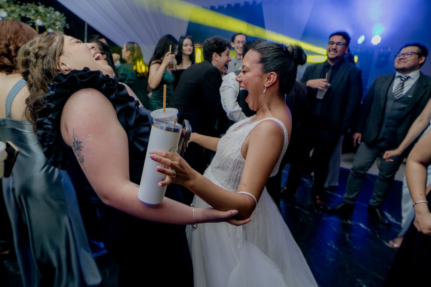 Bride laughing and dancing with guest holding tumbler cup at lively wedding reception party.