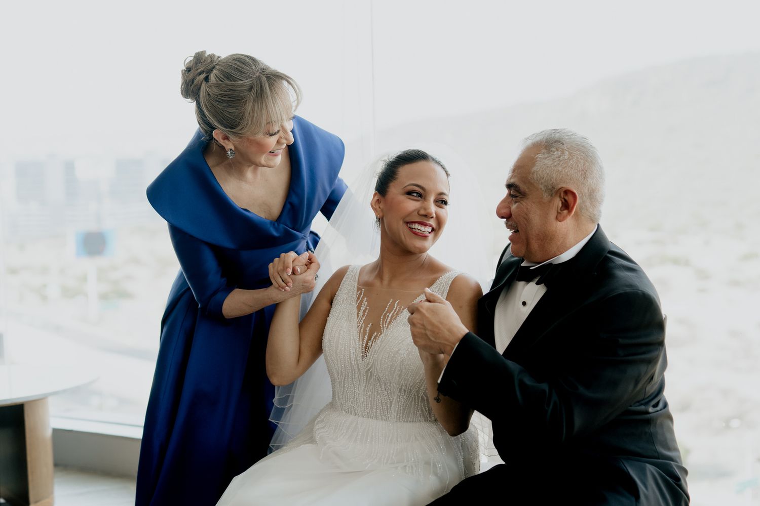 Bride in white gown smiling with parents in formal wedding attire indoors.