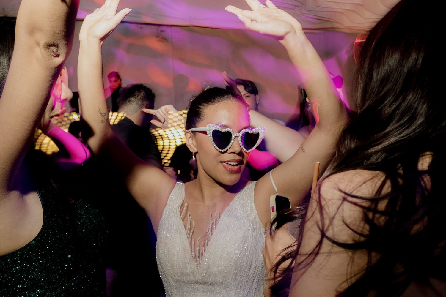 Bride in white dress dances wearing heart-shaped sunglasses at a vibrant wedding reception.