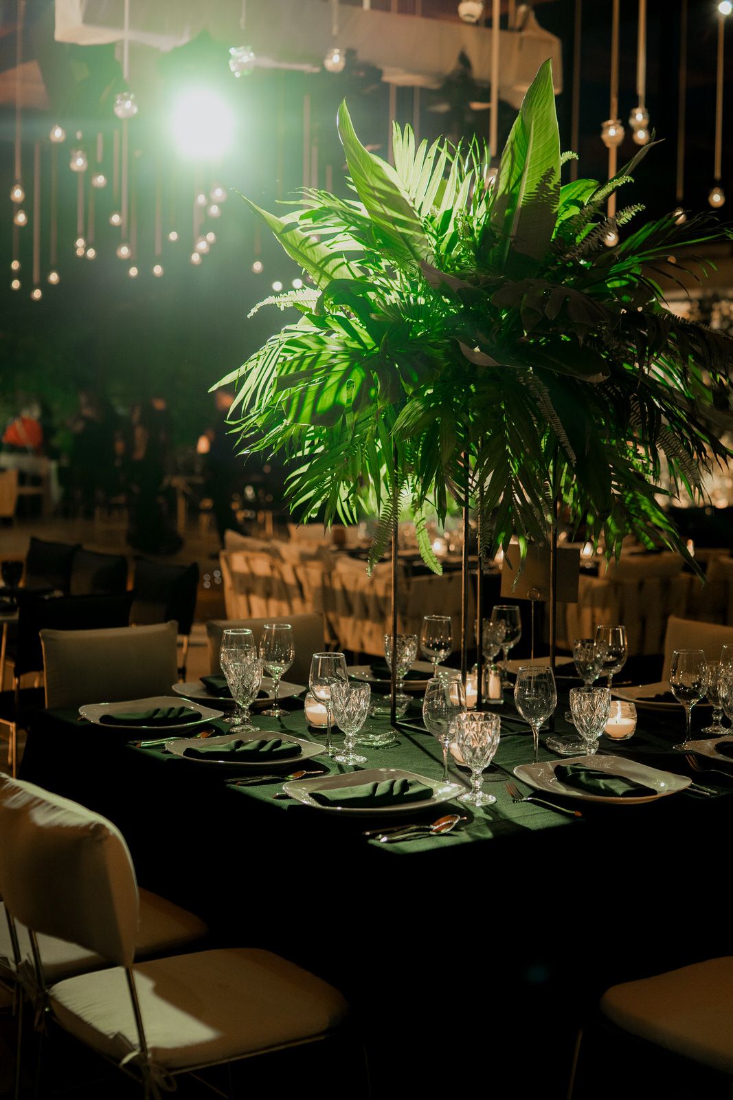 Elegant table settings with palm centerpieces and hanging lights create a tropical ambiance.