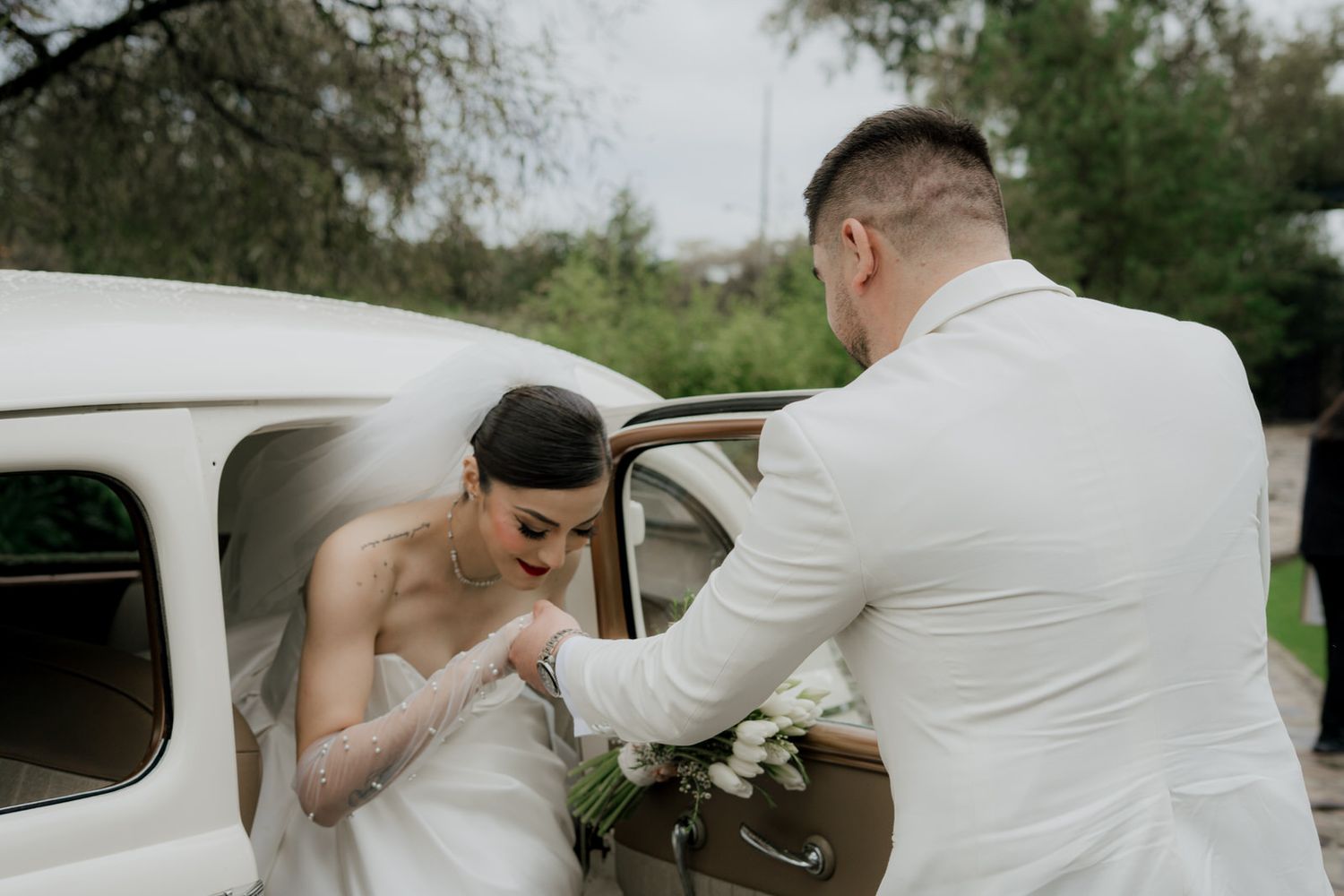 Gentleman assists bride entering vintage white car during wedding photoshoot.