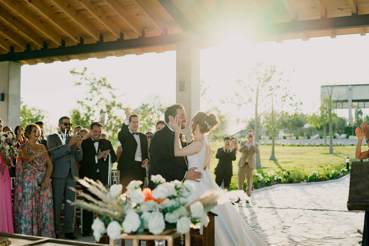 A sun-drenched wedding ceremony under an outdoor pavilion with floral decorations.