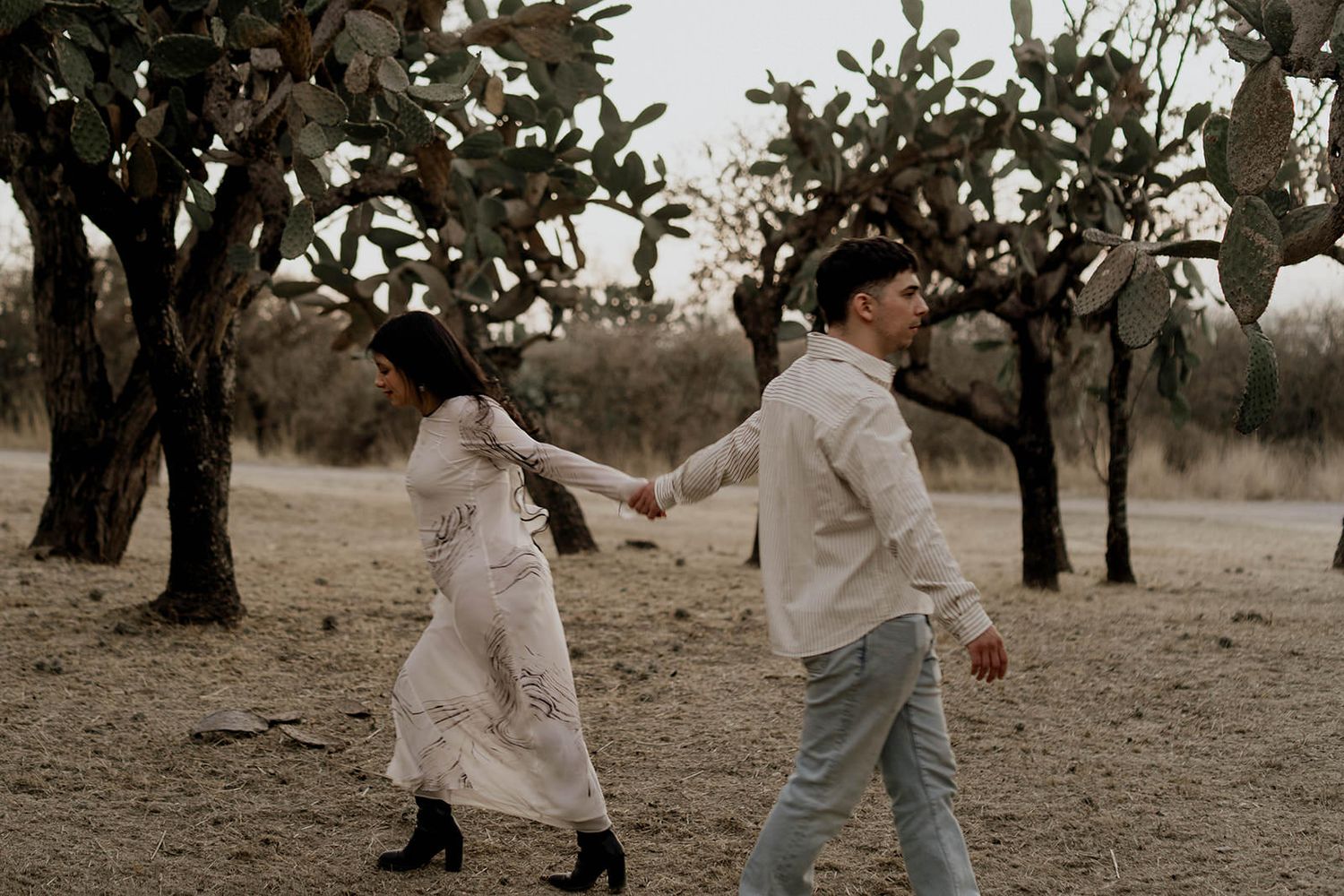 A couple in light colored clothing dances together among desert trees at dusk.