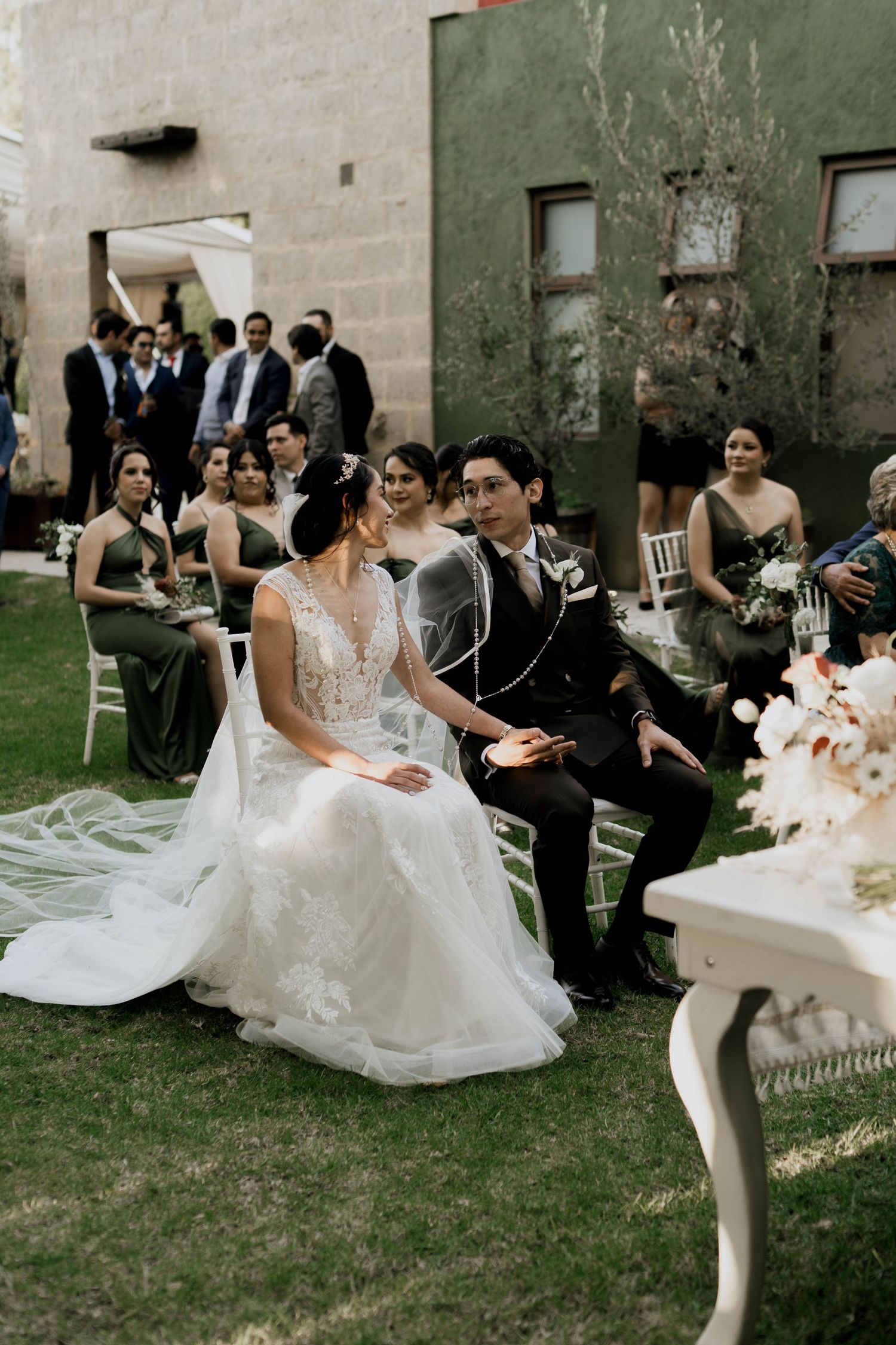 Wedding party seated on white chairs during an outdoor ceremony with floral decorations.