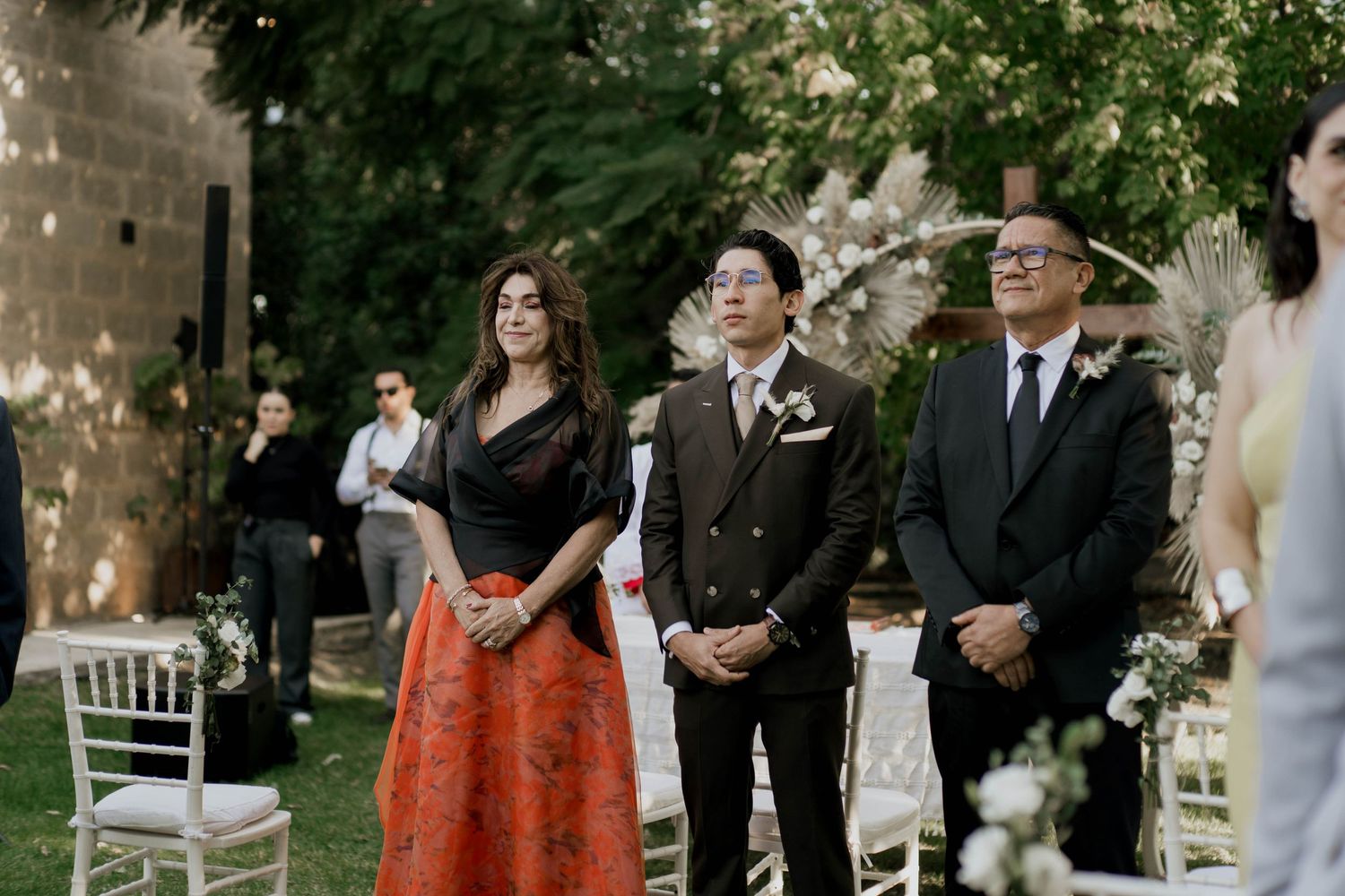 Guests in formal attire stand among white chairs at outdoor garden wedding.
