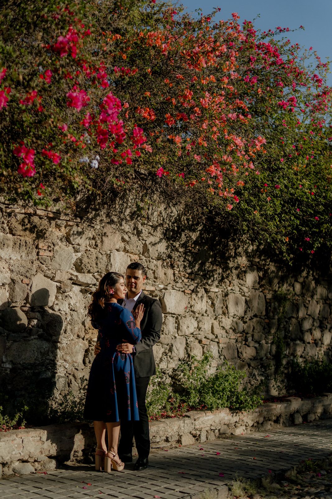 A romantic moment against a stone wall with vibrant pink and orange bougainvillea flowers blooming above.