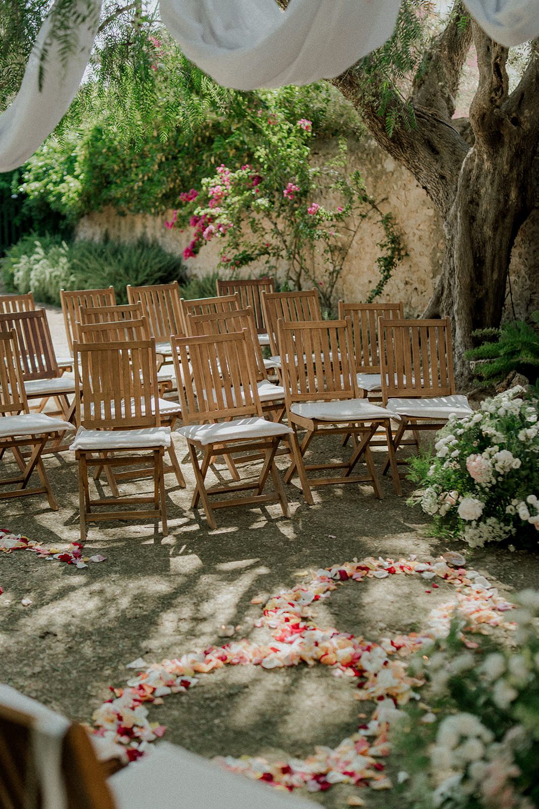 Wooden folding chairs arranged for an outdoor garden wedding ceremony with flower petals scattered on the ground.