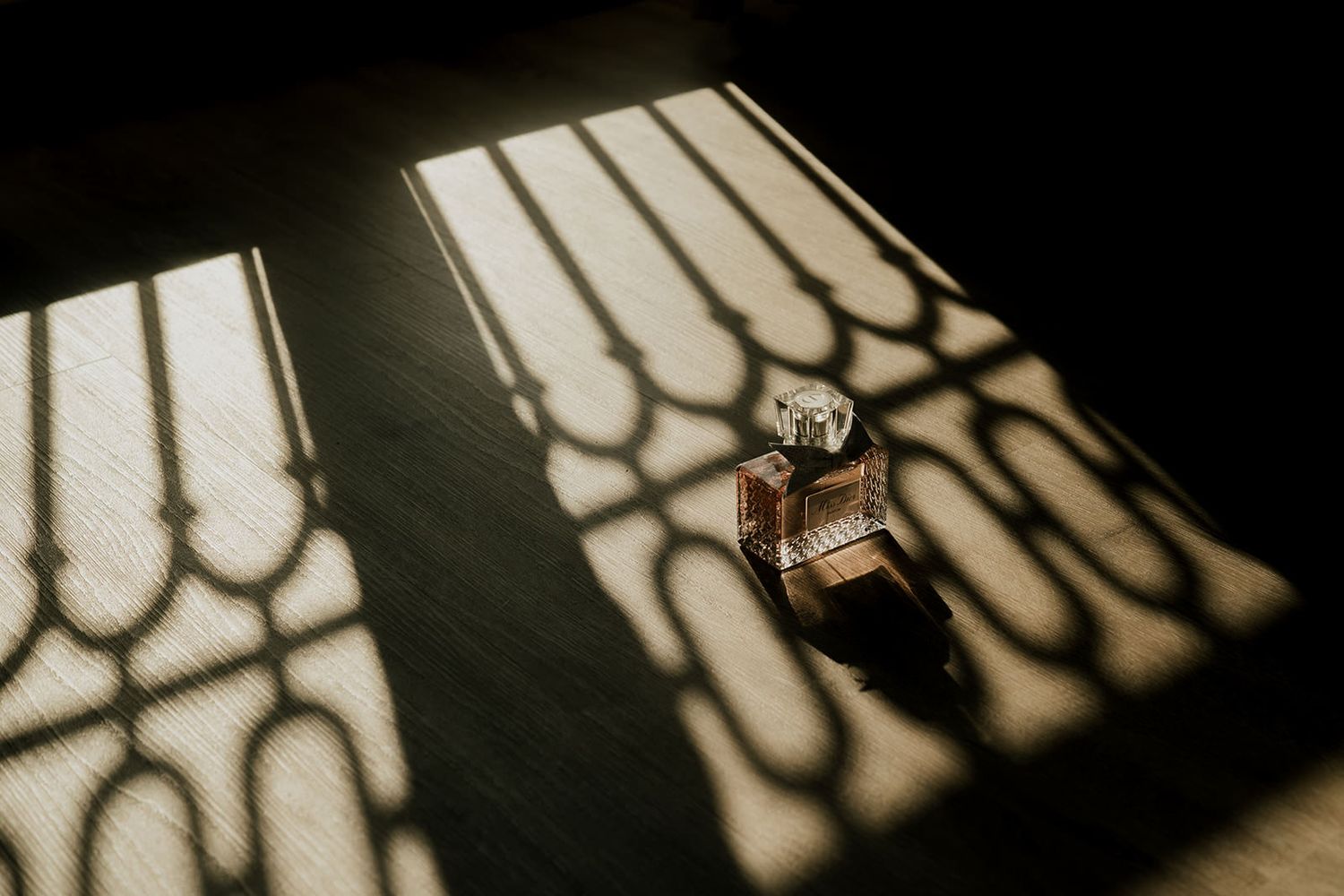 A perfume bottle casting elegant shadows on a dark surface with ornate light patterns from a window.