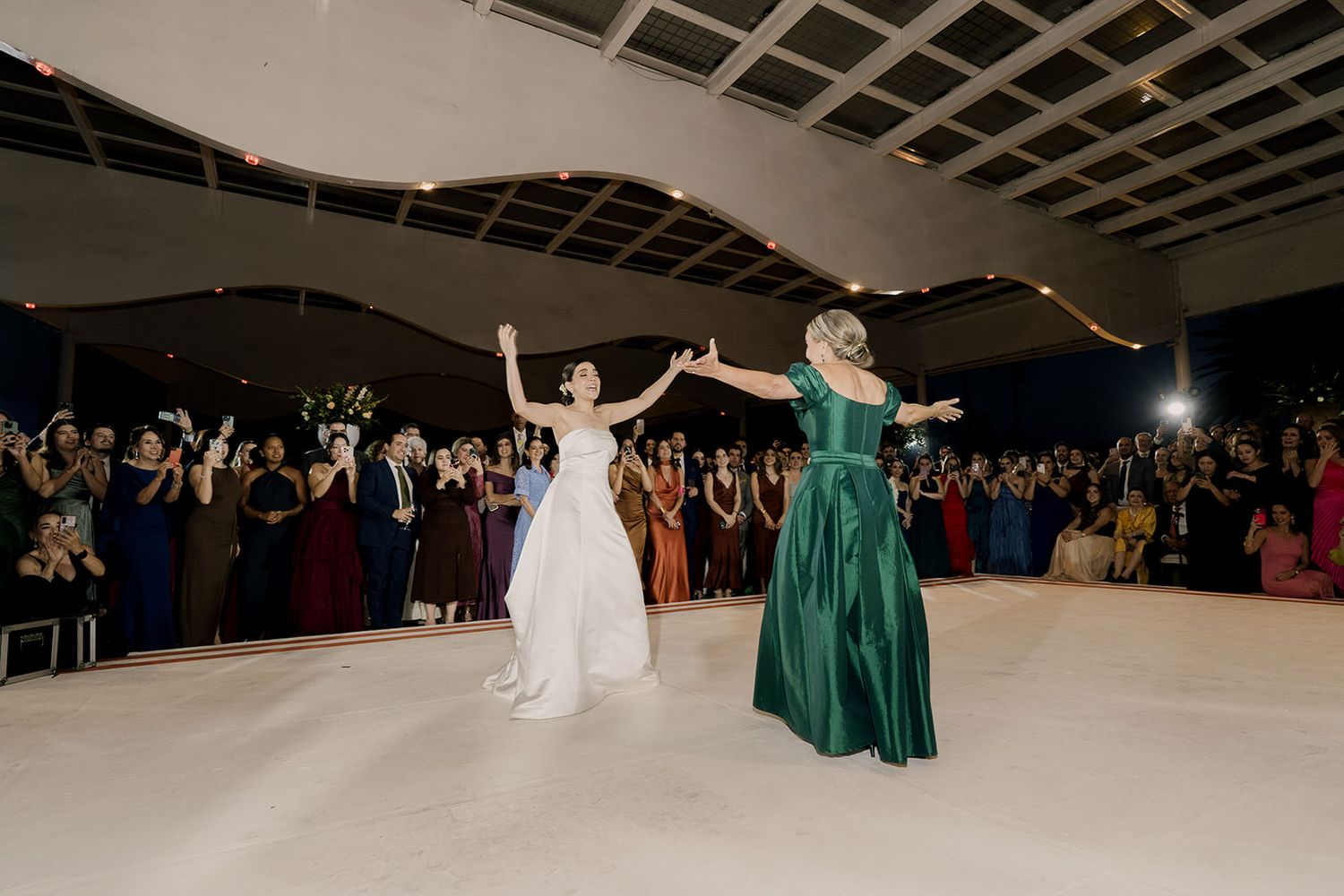 Bride in white gown and woman in green dress celebrate together on dance floor at wedding reception.
