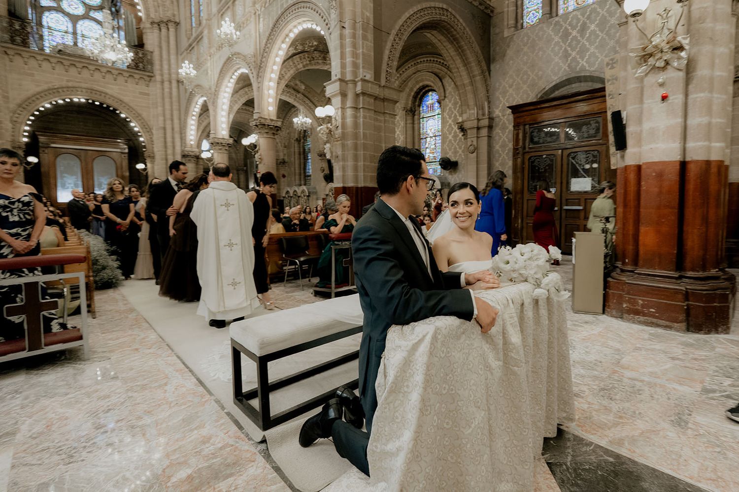 Bride and groom kneeling at altar during elegant church wedding ceremony with guests seated in ornate cathedral.