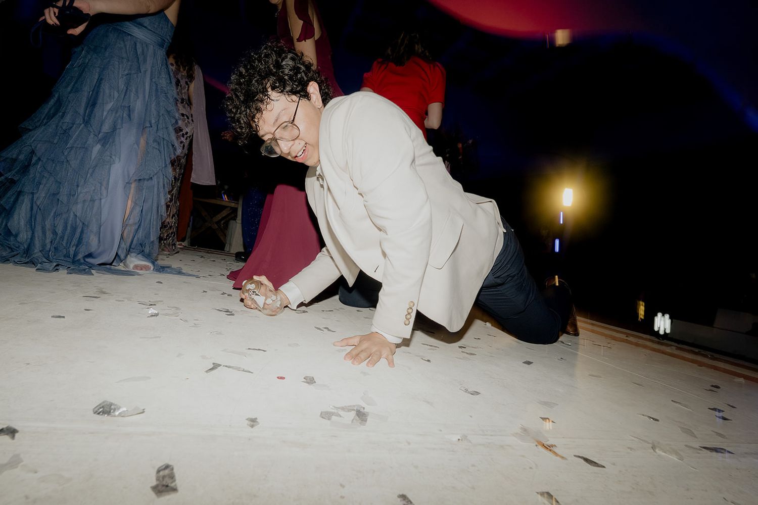 Wedding guest in white blazer playfully crawls on confetti-covered dance floor during lively reception party.