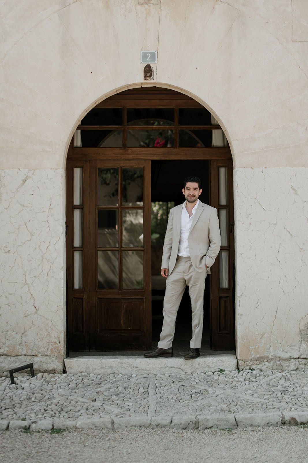 A person in a light colored suit stands in an arched doorway of a rustic stone building.