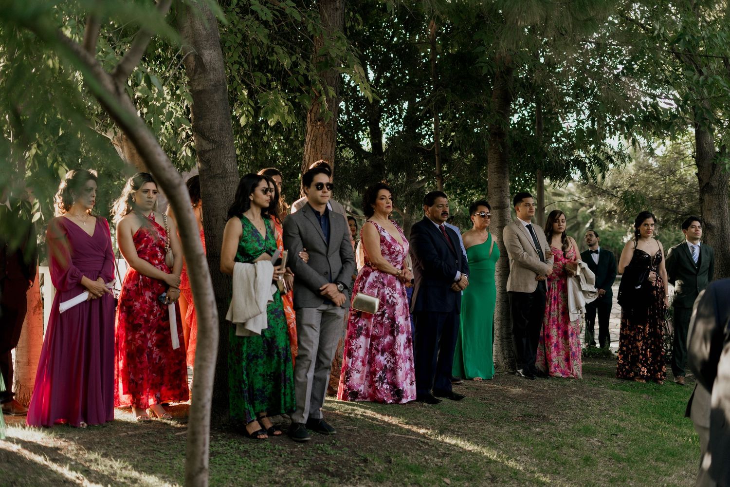 Wedding guests in colorful attire gather under trees for a group photo.