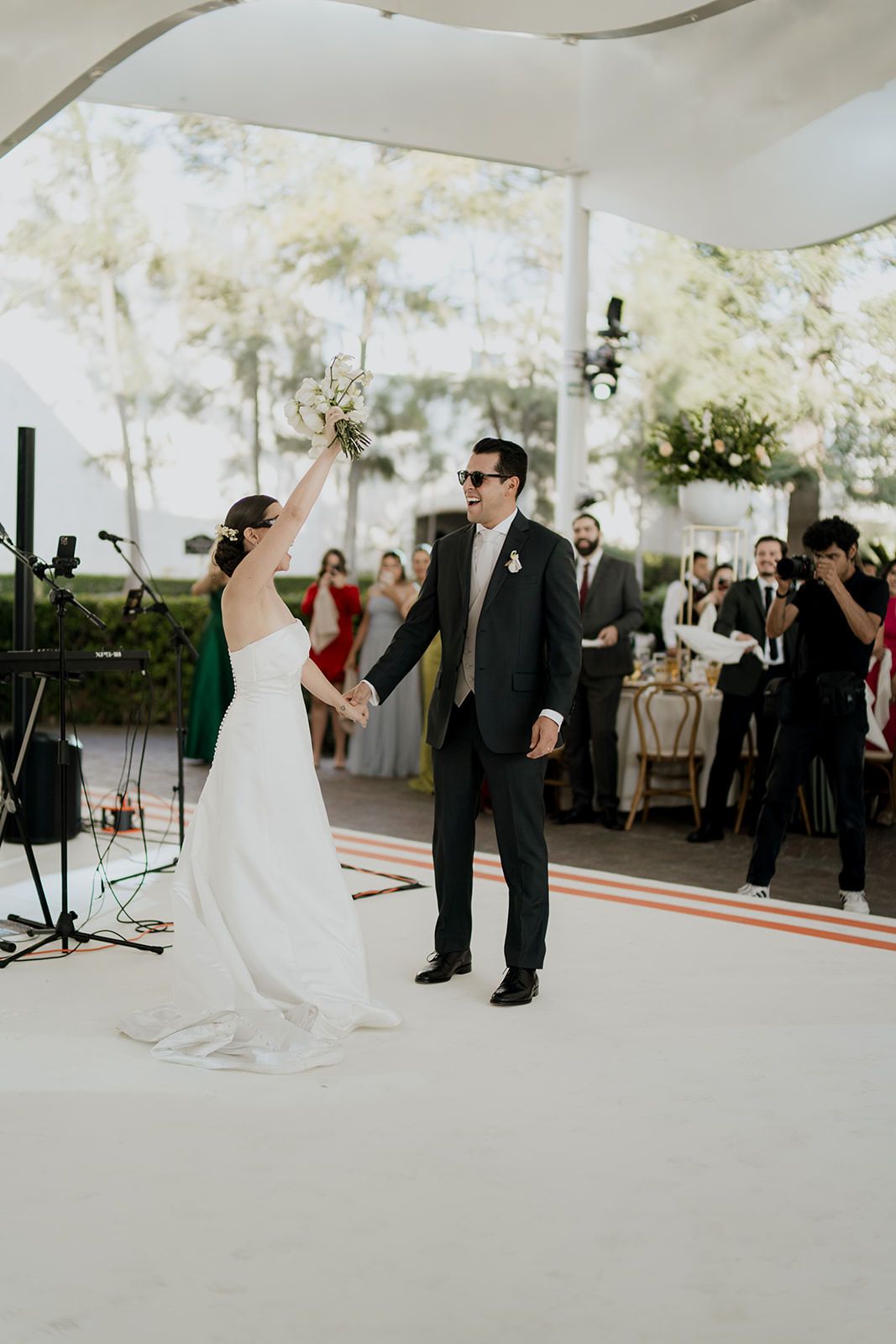 Bride raises bouquet as groom in sunglasses holds her hand at outdoor wedding reception.