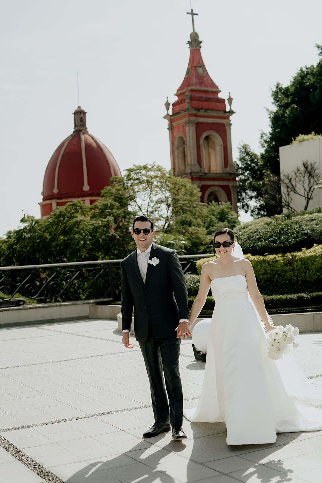 Bride and groom in sunglasses hold hands on terrace with red domed church and bell tower backdrop.