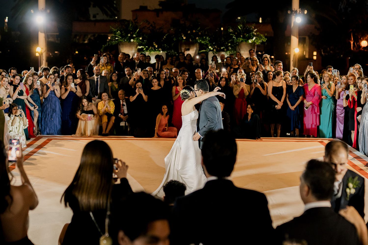Bride and groom share first dance on elegant dance floor surrounded by cheering wedding guests at outdoor evening reception.