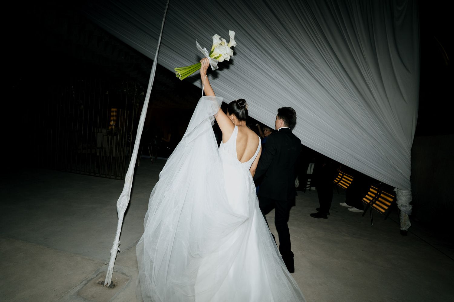 Bride in white gown raises bouquet with groom in black tuxedo at outdoor wedding reception under draped tent.