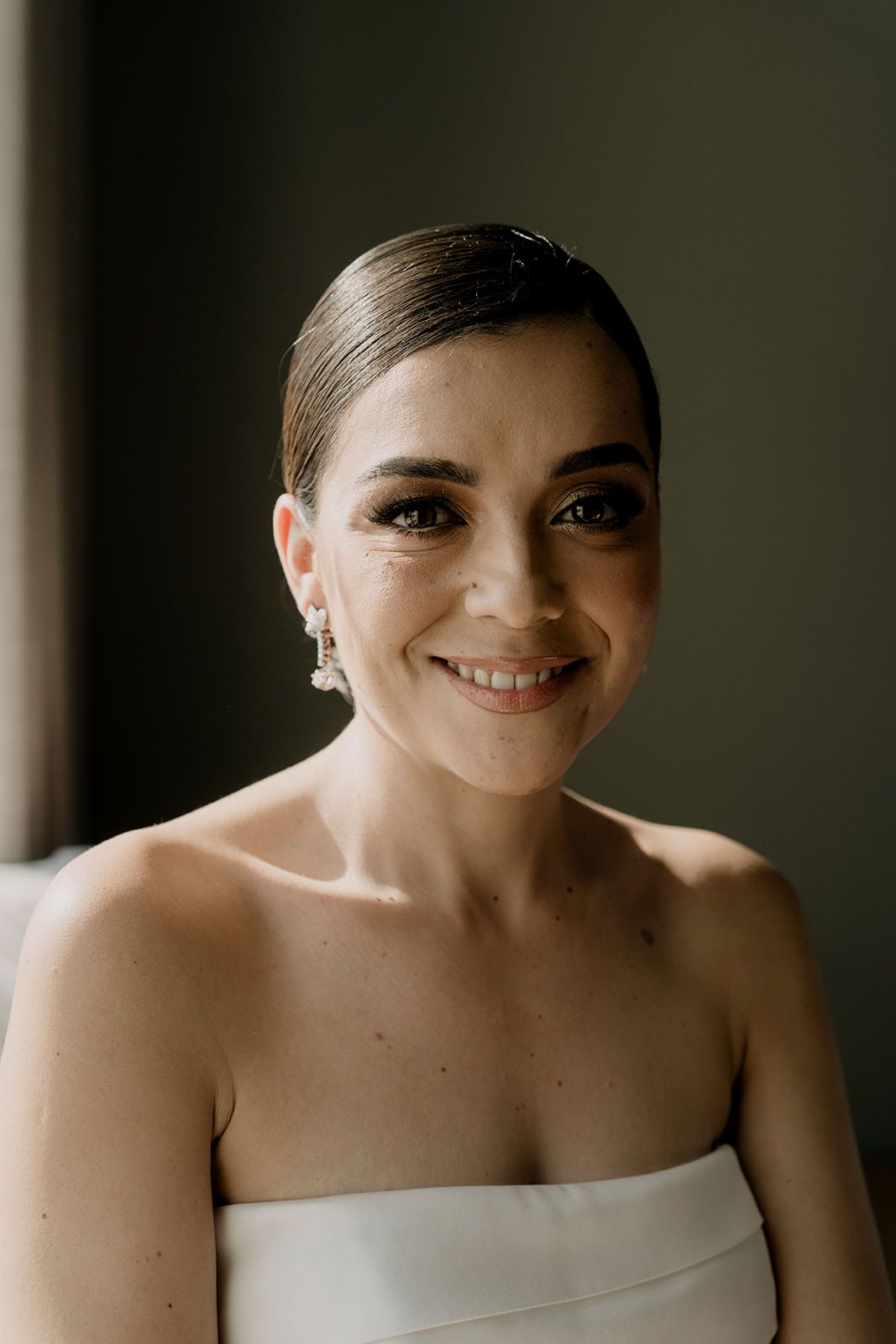 Beautiful bride with sleek updo and pearl earrings smiling in strapless white wedding gown portrait.