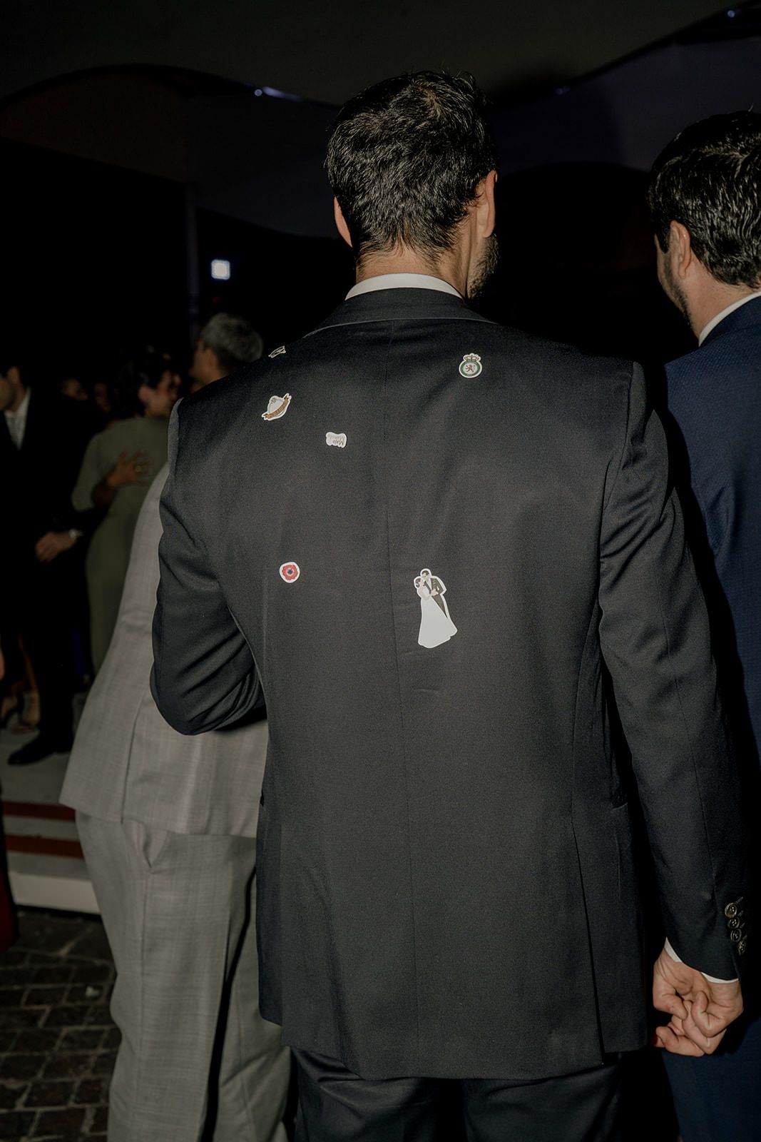 Groom's dark suit jacket decorated with wedding-themed pins and patches at reception.