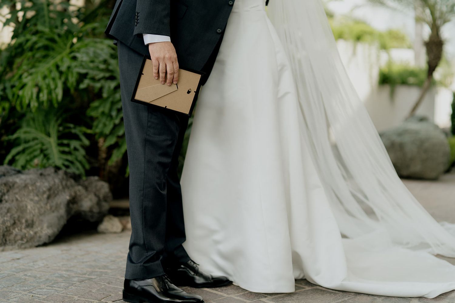 Close-up of wedding couple standing together, groom holding a kraft envelope, bride in elegant white gown with flowing veil.