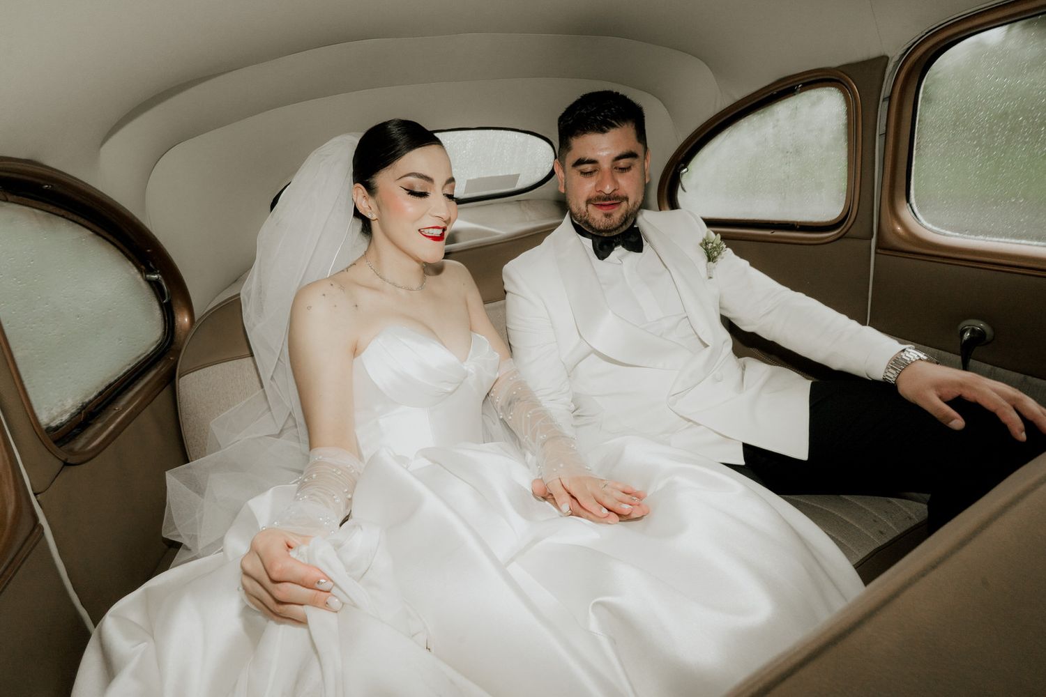 A couple in wedding attire share an intimate moment in the backseat of a vintage car.