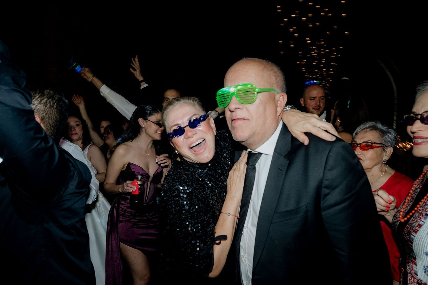 Party guests wearing colorful sunglasses dance together at a lively celebration.
