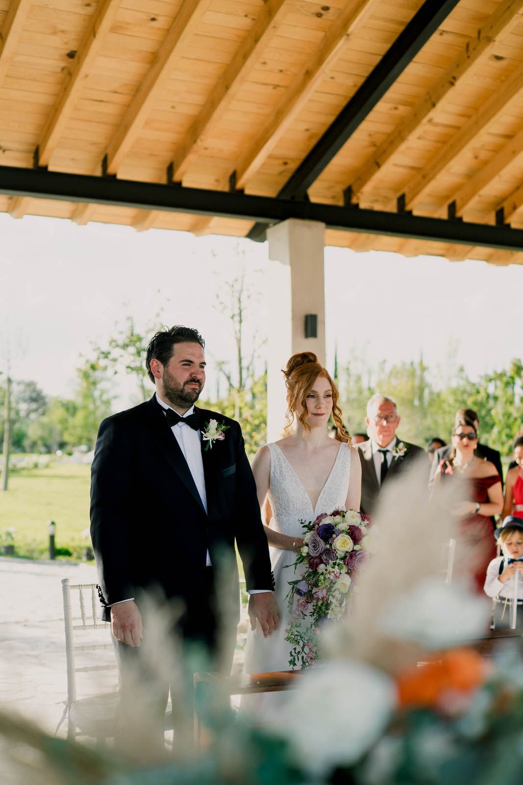 Wedding ceremony taking place under a rustic wooden pergola with natural lighting.