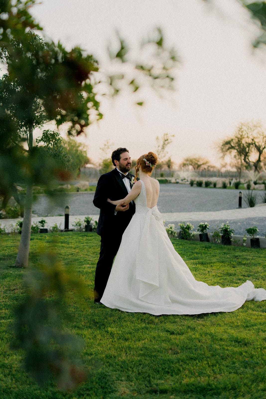 A romantic wedding couple embraces on a manicured lawn at sunset with trees and mountains in the background.