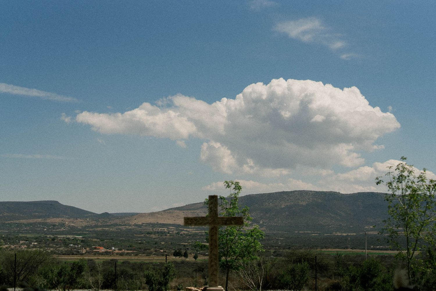 Wooden cross stands against mountain landscape with white puffy clouds in blue sky.
