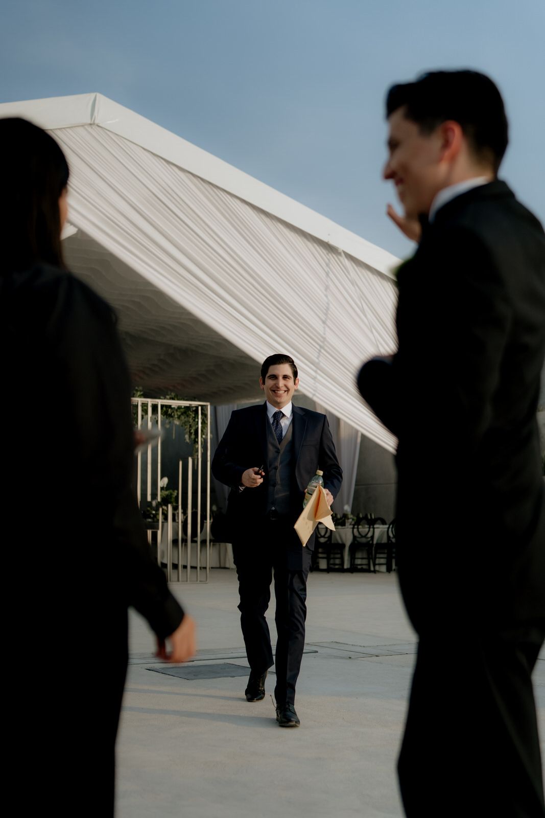 Smiling man in dark suit walks toward camera outside elegant white draped wedding venue tent at golden hour.