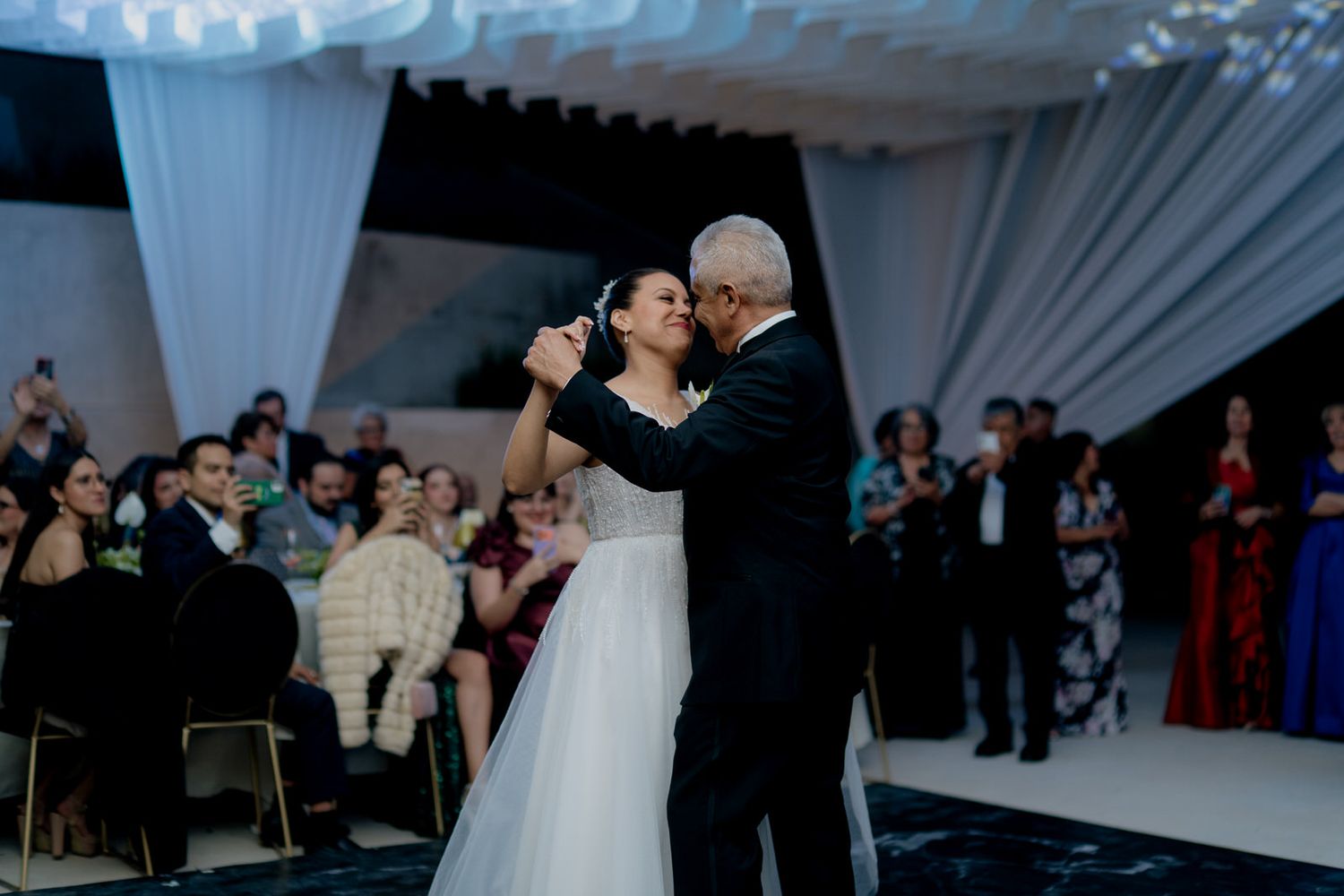 Bride and groom share their first dance at elegant wedding reception with guests watching in background.