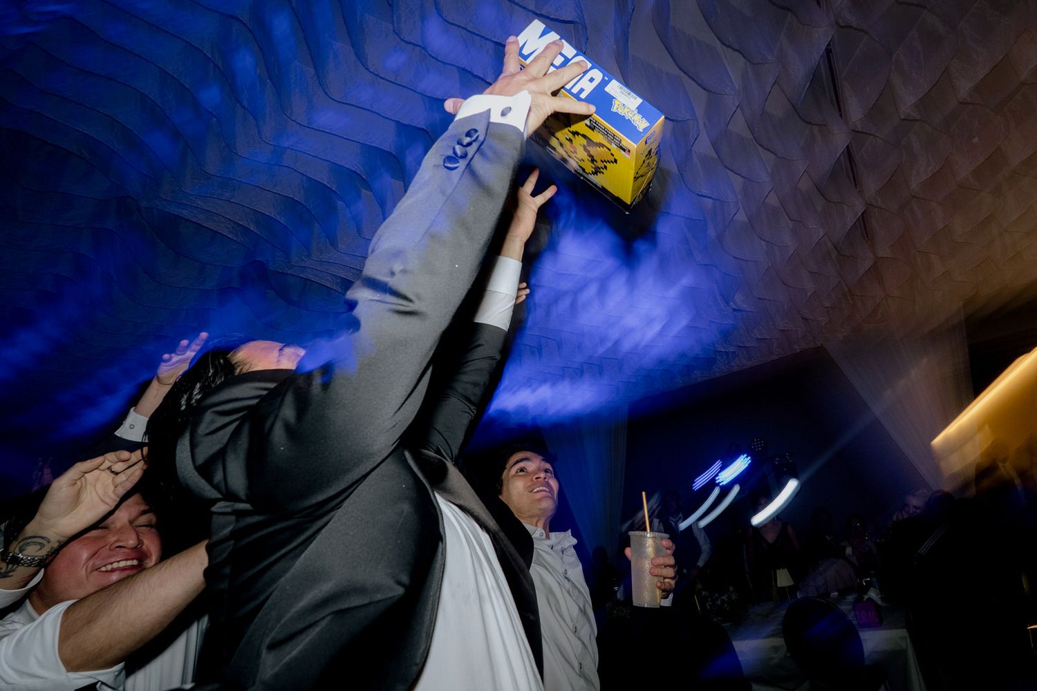 Party guests celebrate at a nightclub, one person raising a beer box overhead under blue ambient lighting.