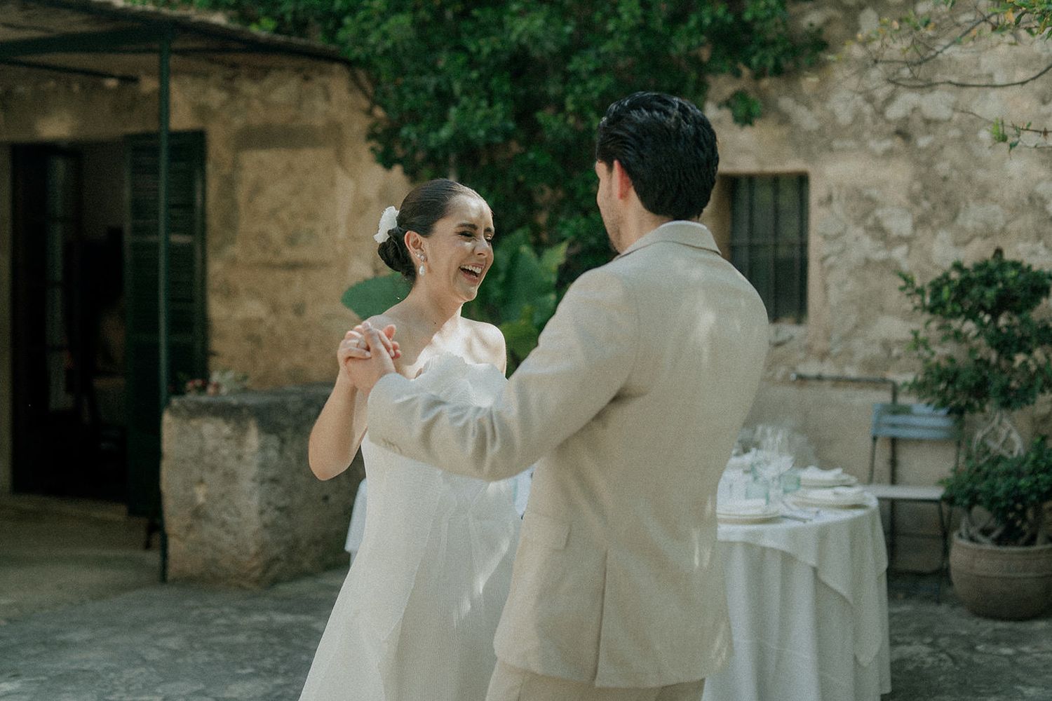 A romantic couple in elegant wedding attire dances together in front of a rustic stone building.