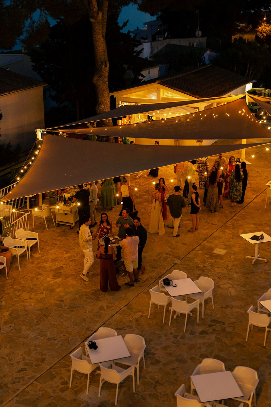 Outdoor evening event setup with white tables and chairs under string lights on a sandy terrace.