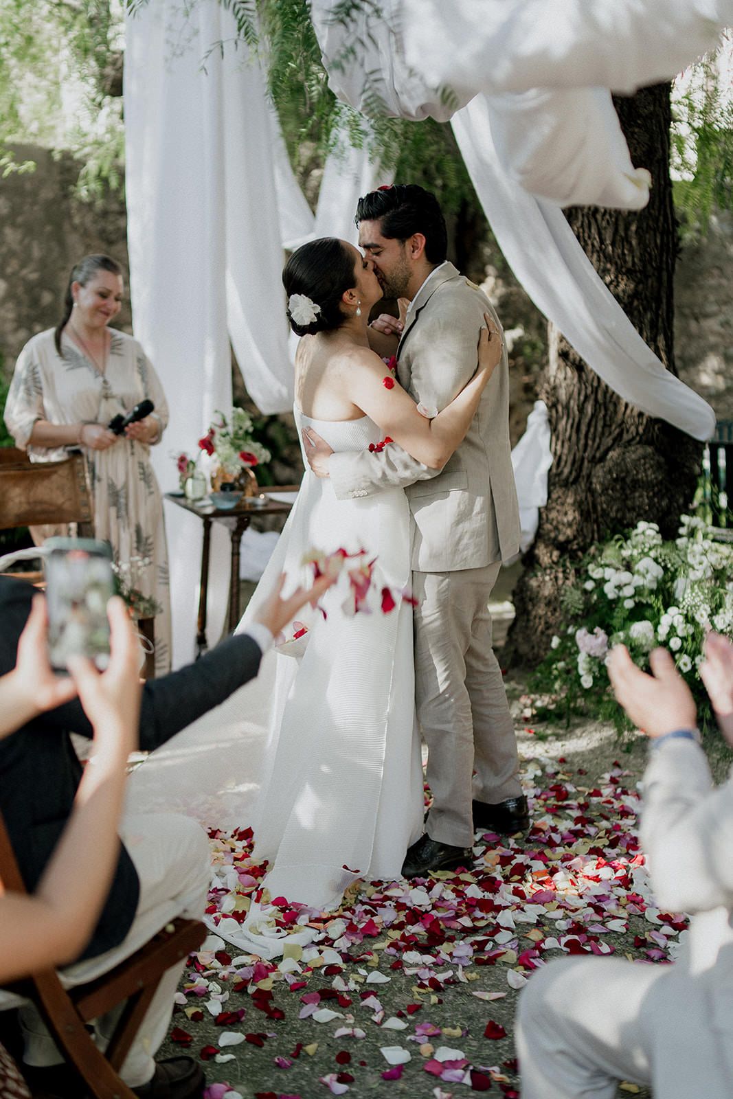 A romantic outdoor wedding ceremony under white draped fabric with rose petals scattered on the ground.
