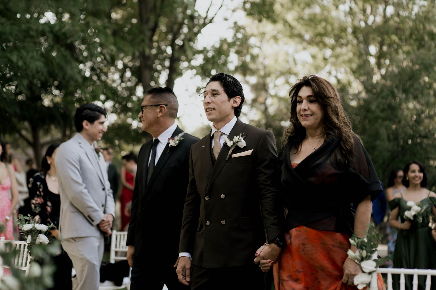 Wedding guests in formal attire stand among white chairs at an outdoor ceremony.