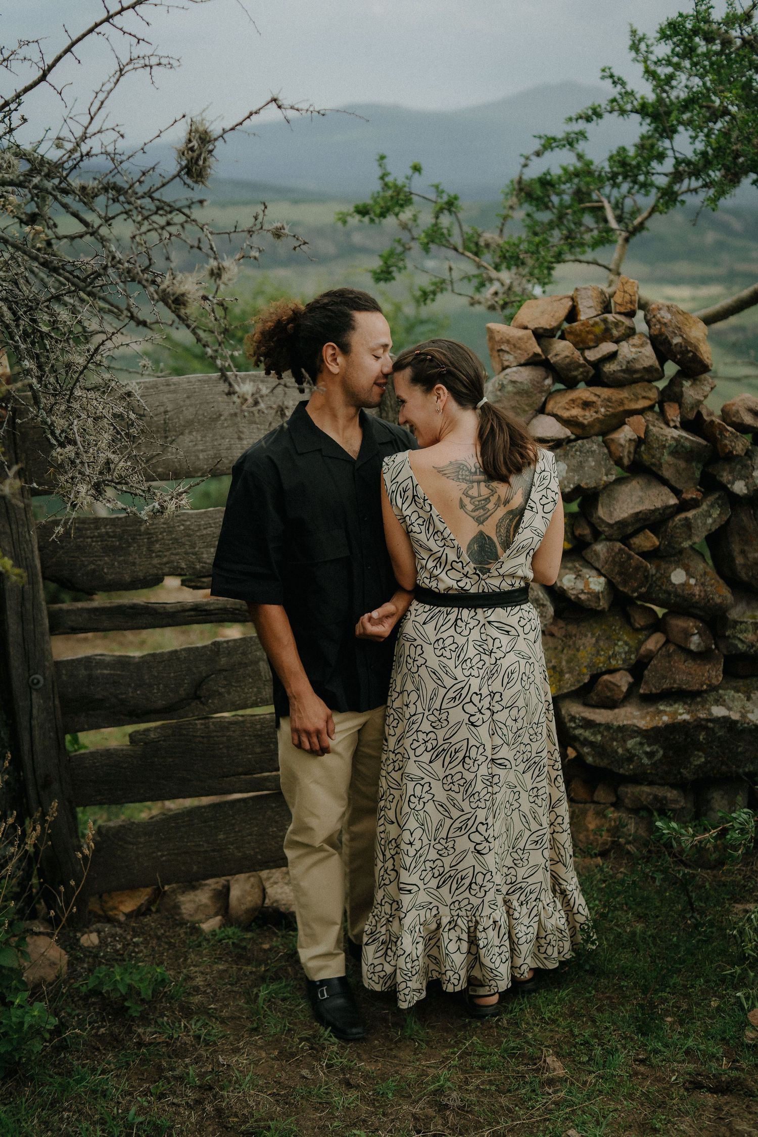 A romantic couple shares an intimate moment by a rustic stone wall in a mountainous countryside setting.