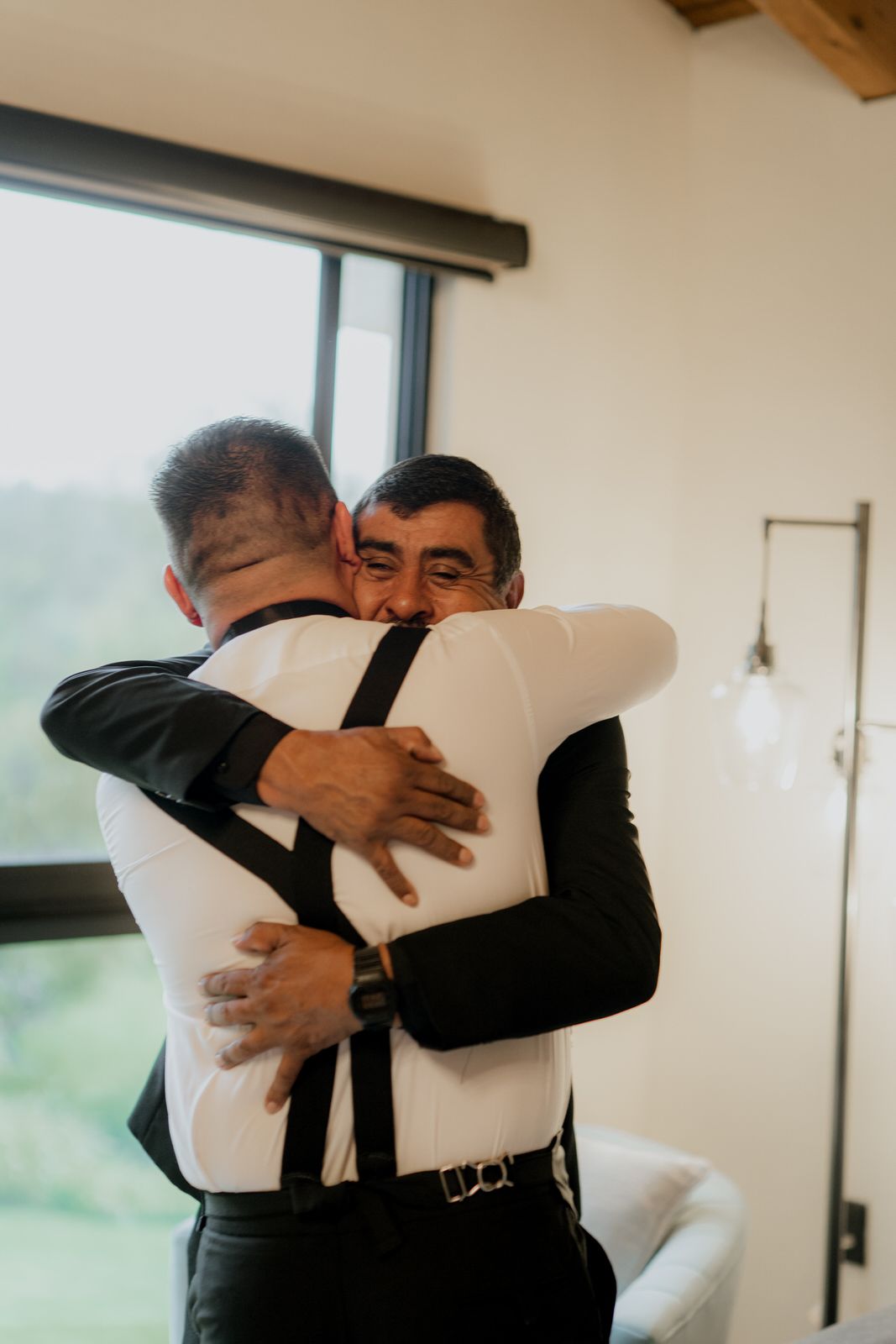 Emotional group hug between wedding party members in a bright room with large windows.