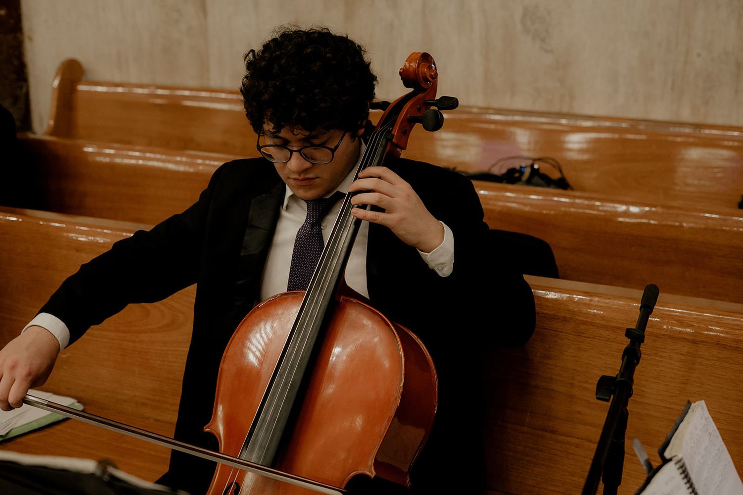 Young cellist in black suit playing cello while reading sheet music inside a church at a wedding.