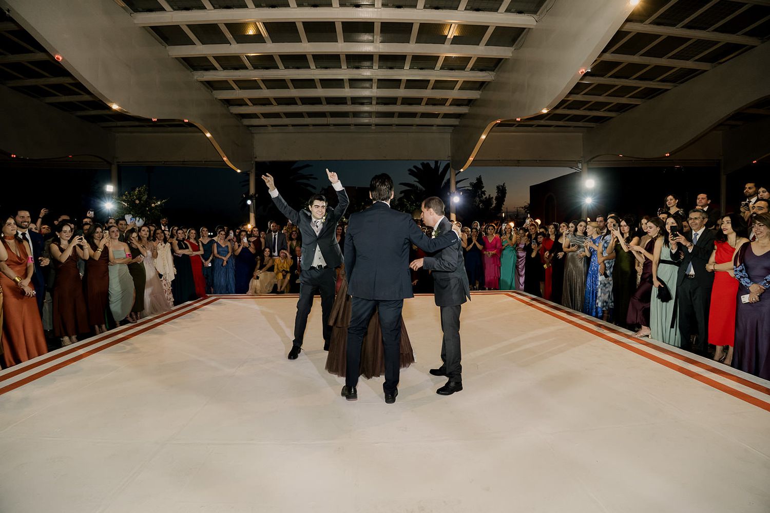 Bride and groomsmen celebrate on white dance floor as wedding guests cheer and applaud at night reception.