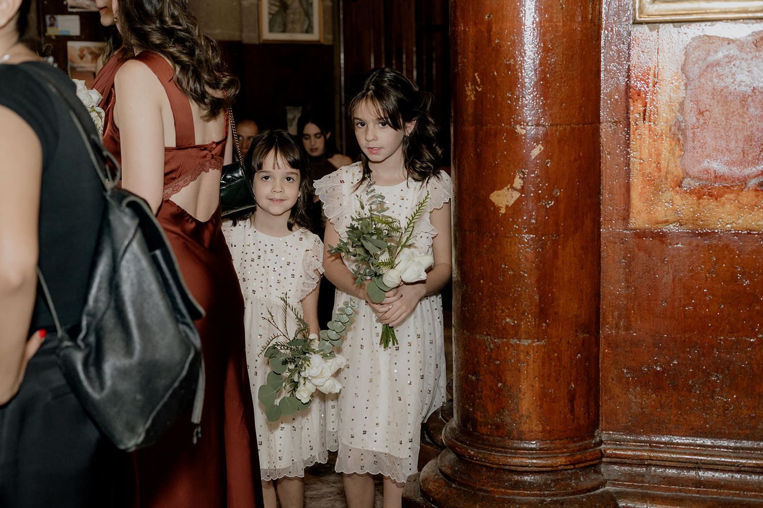 Two young flower girls in white dresses holding bouquets of white flowers and greenery at a wedding ceremony.