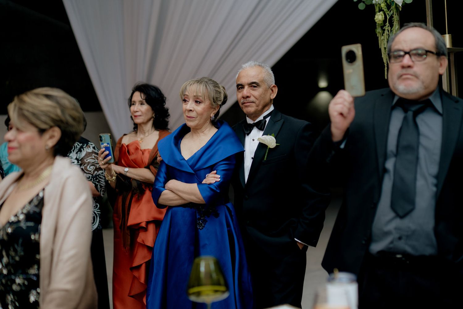 Elegantly dressed wedding guests in formal attire, including a woman in blue gown and man in tuxedo, watching a ceremony.