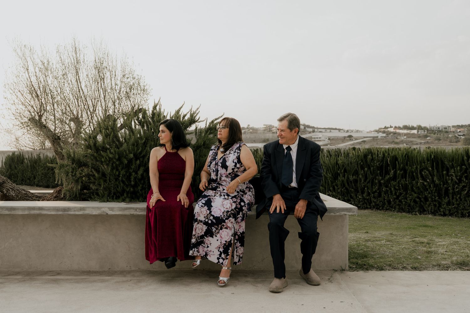 Three people sitting on a stone wall outdoors, dressed in formal attire, with rolling hills in the background.