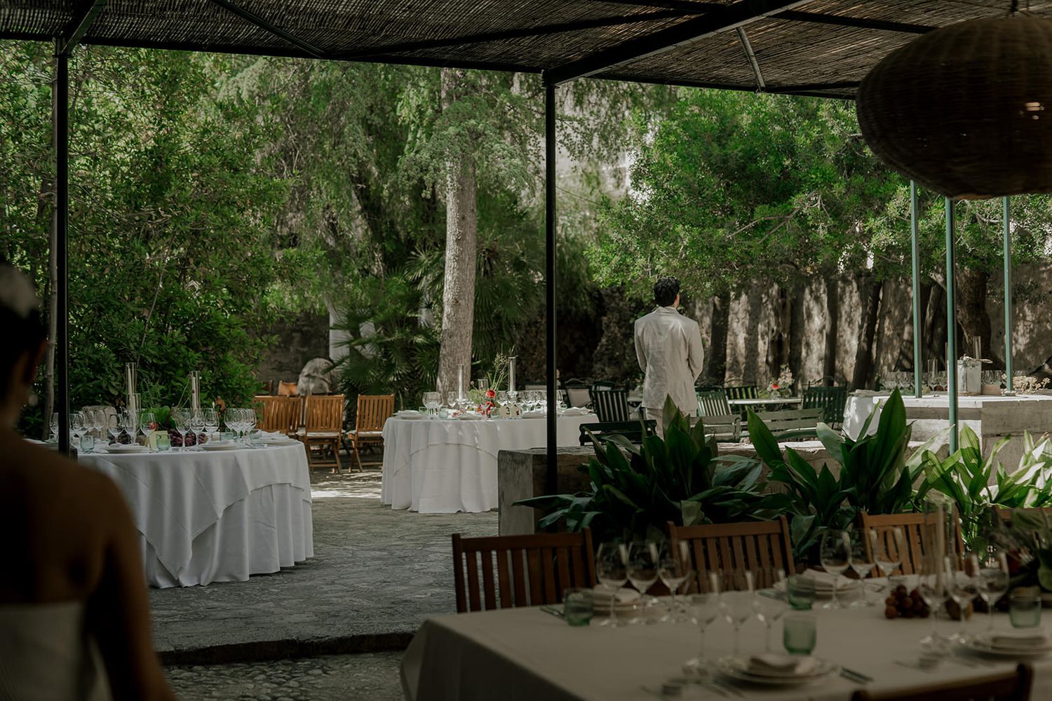 Outdoor wedding reception setup with wooden chairs and tables under natural tree canopy.