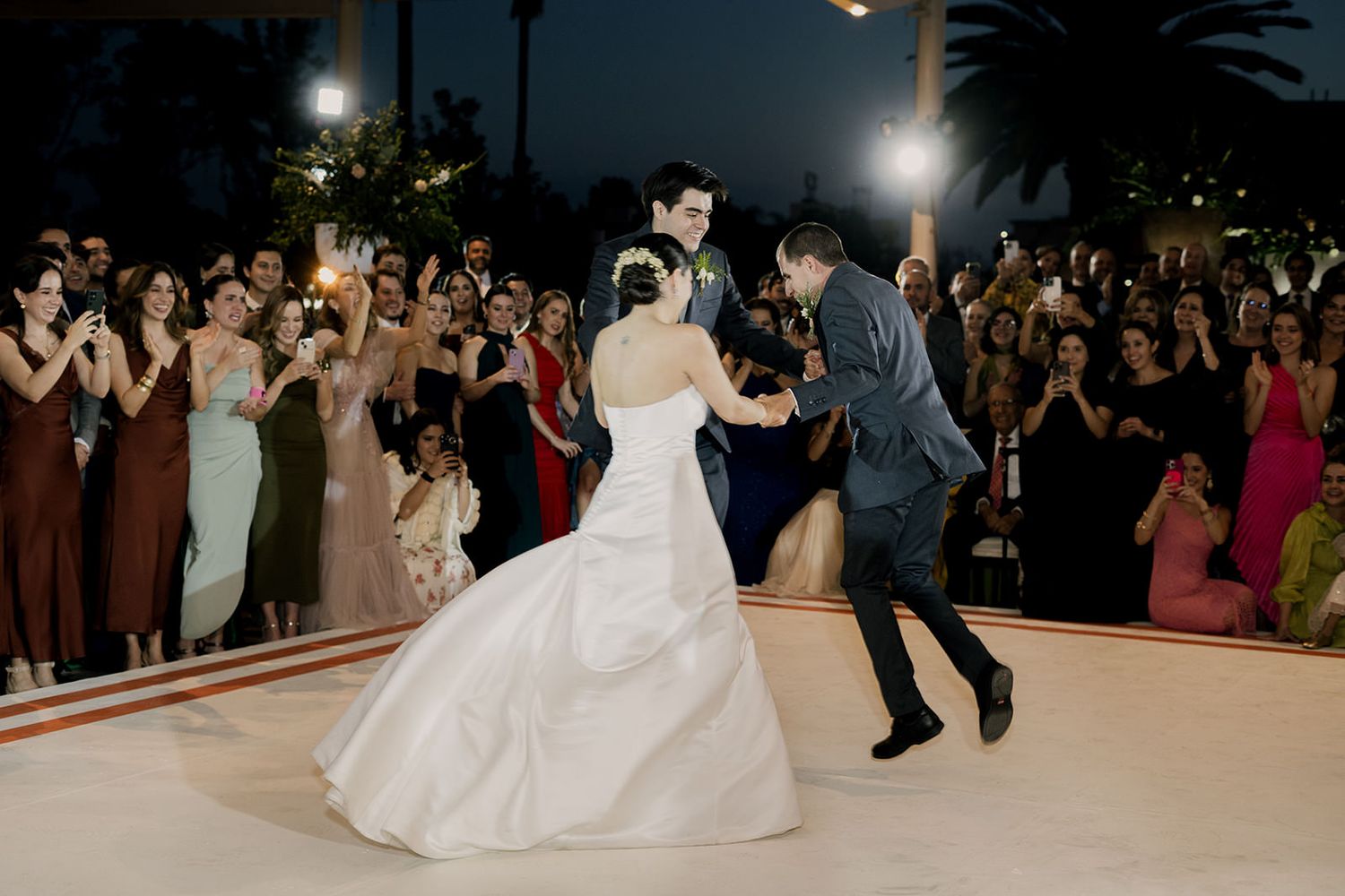 Bride and groom share romantic first dance on white dance floor surrounded by cheering wedding guests at night.