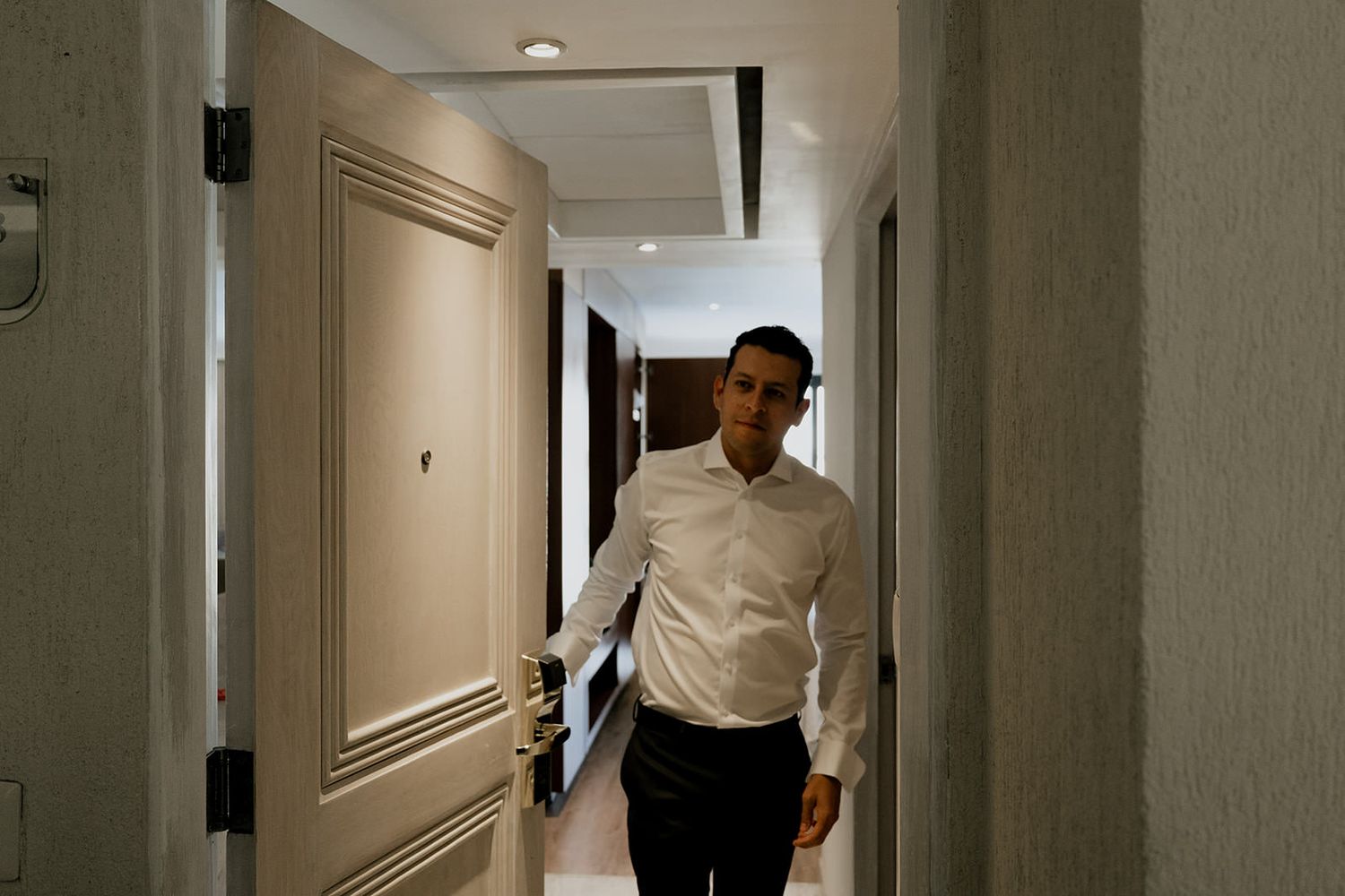 Man in white dress shirt walking through a modern hotel corridor with beige walls and doors.