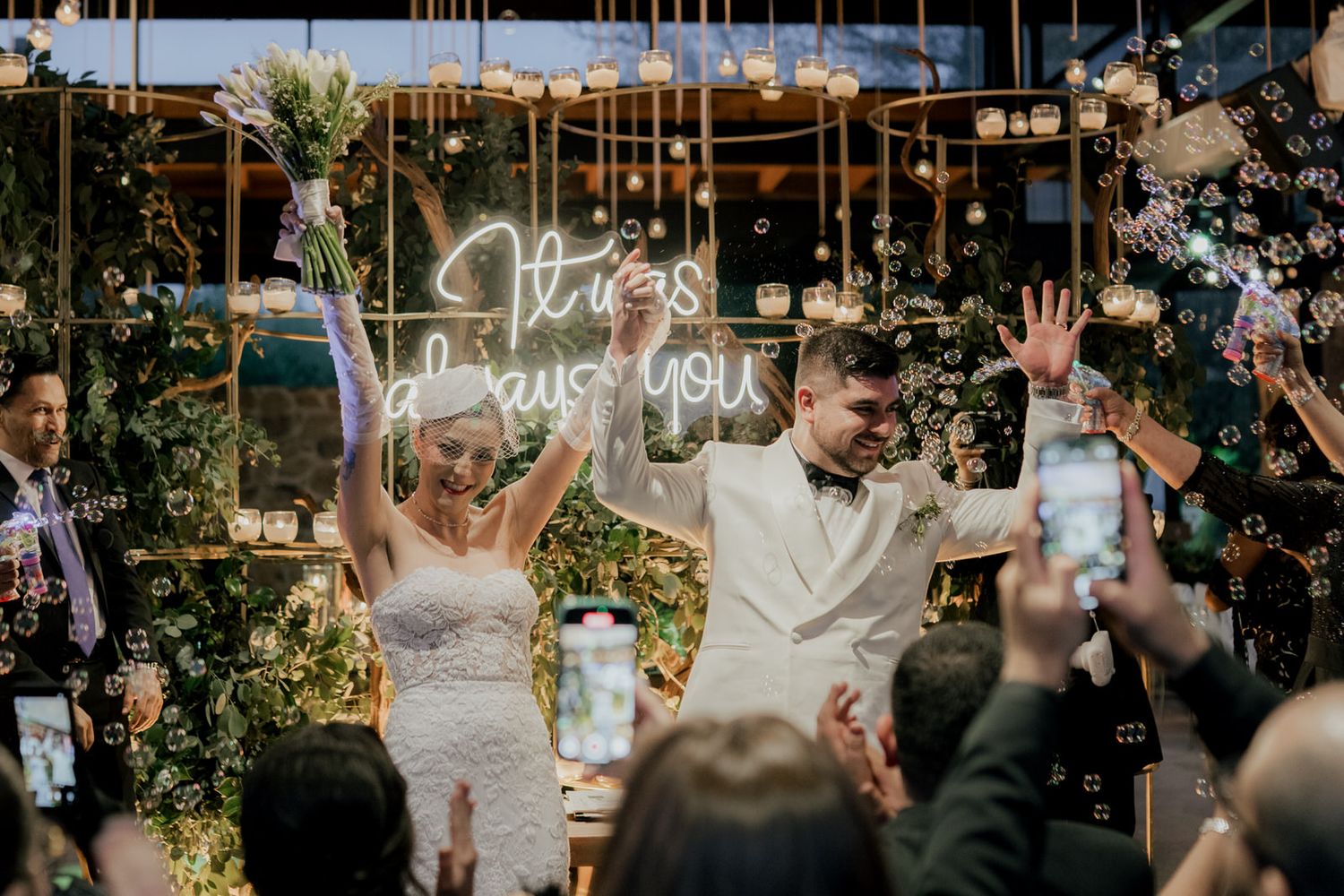 A couple celebrates with raised arms under string lights and floral decorations at their reception.